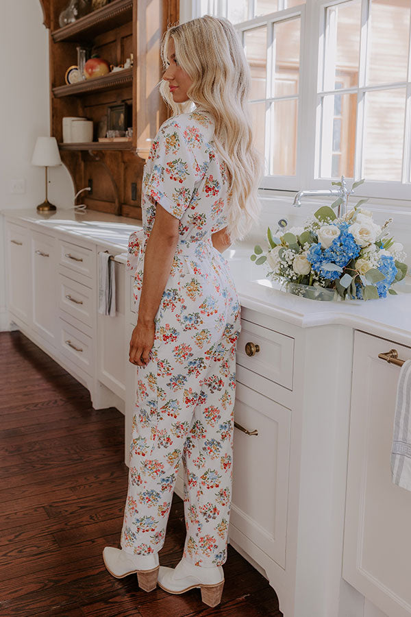 Woman poses in a charming white floral jumpsuit with tailored details, accessorized with sleek white boots for a winter-chic aesthetic.